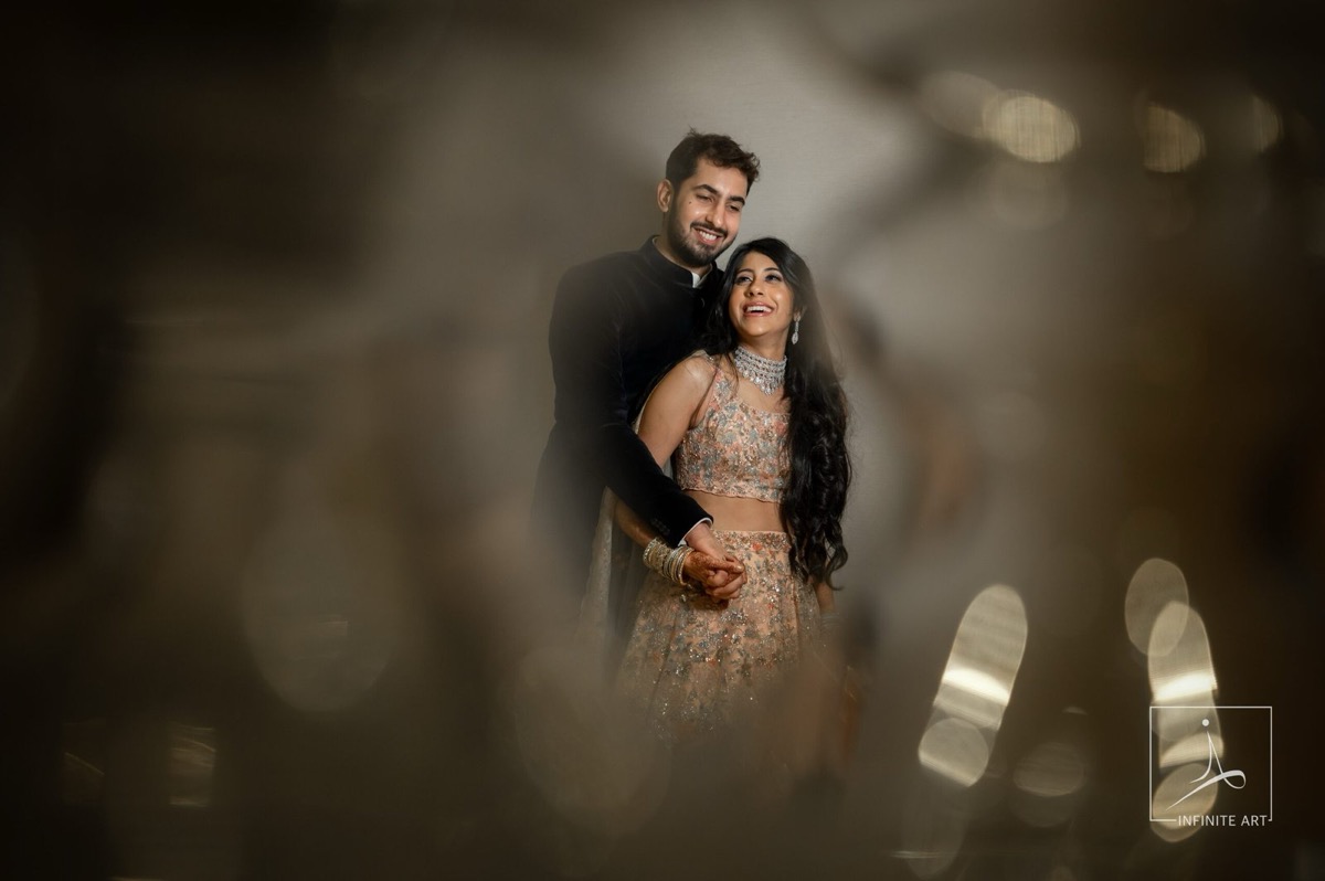 Abhishek and Shweta smiling, framed through soft bokeh — he in a dark bandhgala, she in an embroidered peach lehenga