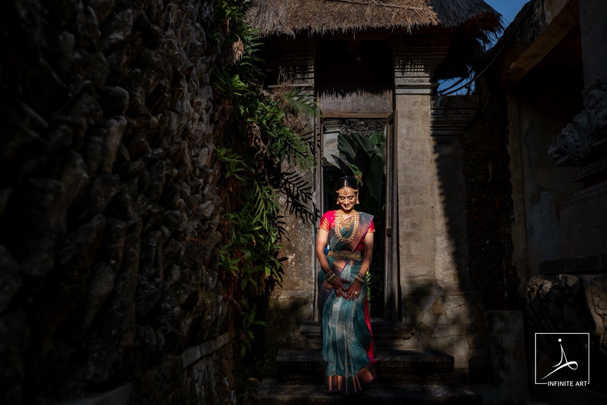 A bride in a teal and pink silk saree stands on stone steps in a sunlit doorway beneath a thatched roof