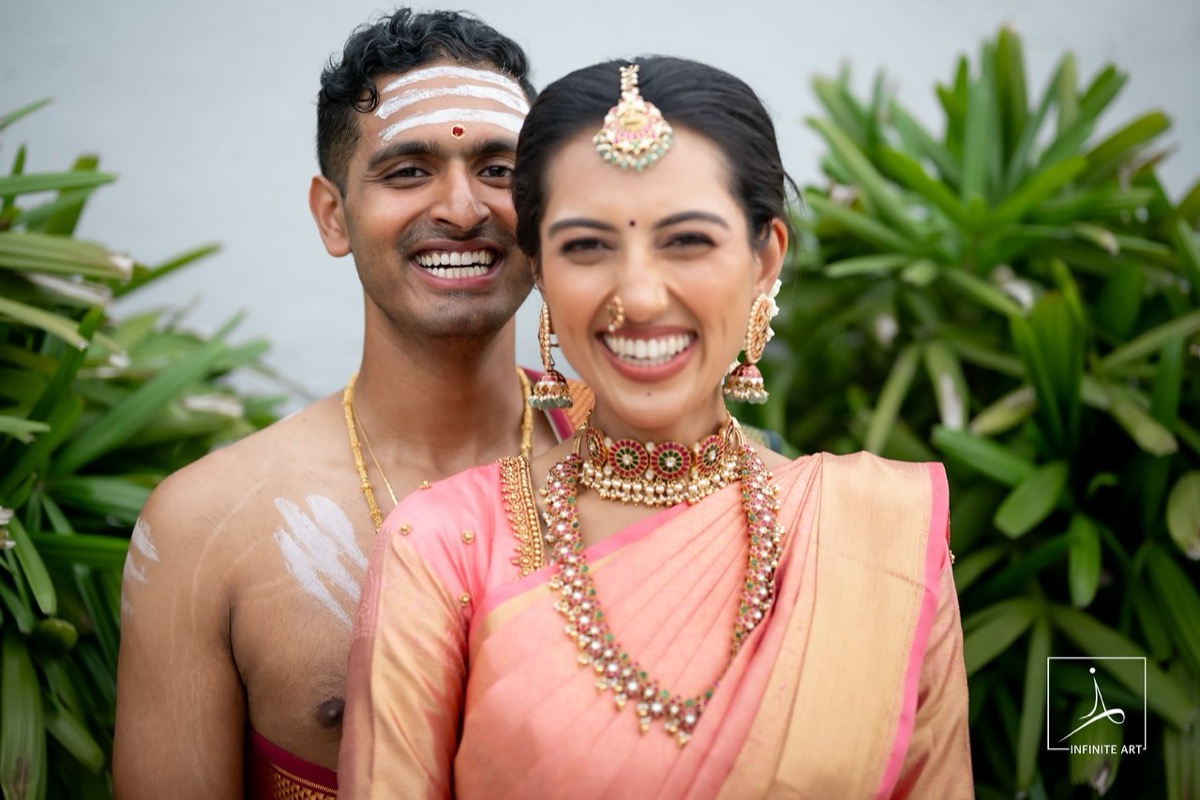 A groom with sacred ash on his forehead and a bride in a pink silk saree and temple jewellery laugh together in front of green leaves