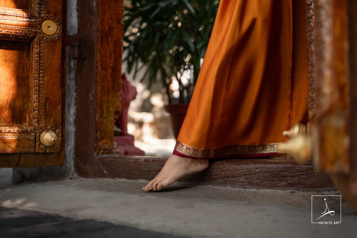 A bride's bare foot steps over a carved wooden threshold, the hem of her orange silk saree lifted, in warm afternoon light